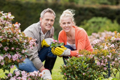A man and woman are smiling and kneeling in a well-maintained garden with vibrant pink and white flowering bushes in the foreground. They are dressed casually for gardening, with the man wearing a light grey shirt and the woman in an orange cardigan, both wearing yellow gardening gloves. The woman is holding a small potted plant, indicating plant care or planting activity. Behind them, the garden features a lush, green lawn and a backdrop of trees and shrubs, suggesting a landscaped outdoor space typical of residential gardens in Finchley. The scene appears to be during daylight hours with natural lighting, possibly on a mild and clear day, reflecting regular outdoor maintenance or gardening services. This image illustrates active gardening work, aligning with services focused on lawn care, shrub trimming, and garden planting for local properties serviced by Gardening Services Finchley.