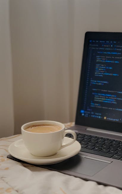 A white ceramic cup filled with coffee resting on a matching saucer sits on a soft, light-colored bedsheet next to an open laptop displaying lines of code on a dark background. The scene is softly lit